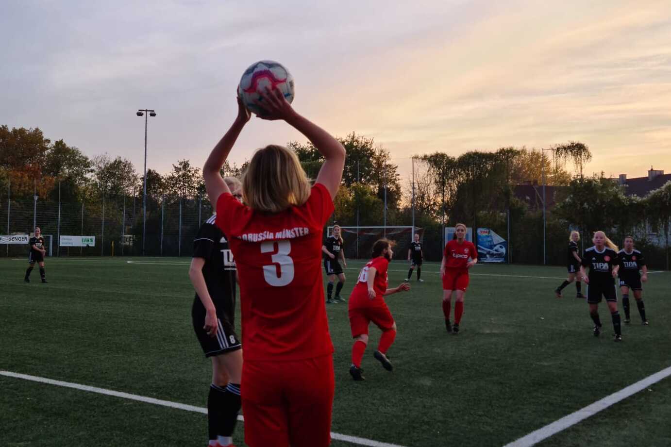 Spielerin in rotem Trikot f&uuml;hrt Einwurf mit Ball auf einem Fu&szlig;ballfeld bei Sonnenuntergang aus, andere Spieler in der N&auml;he.