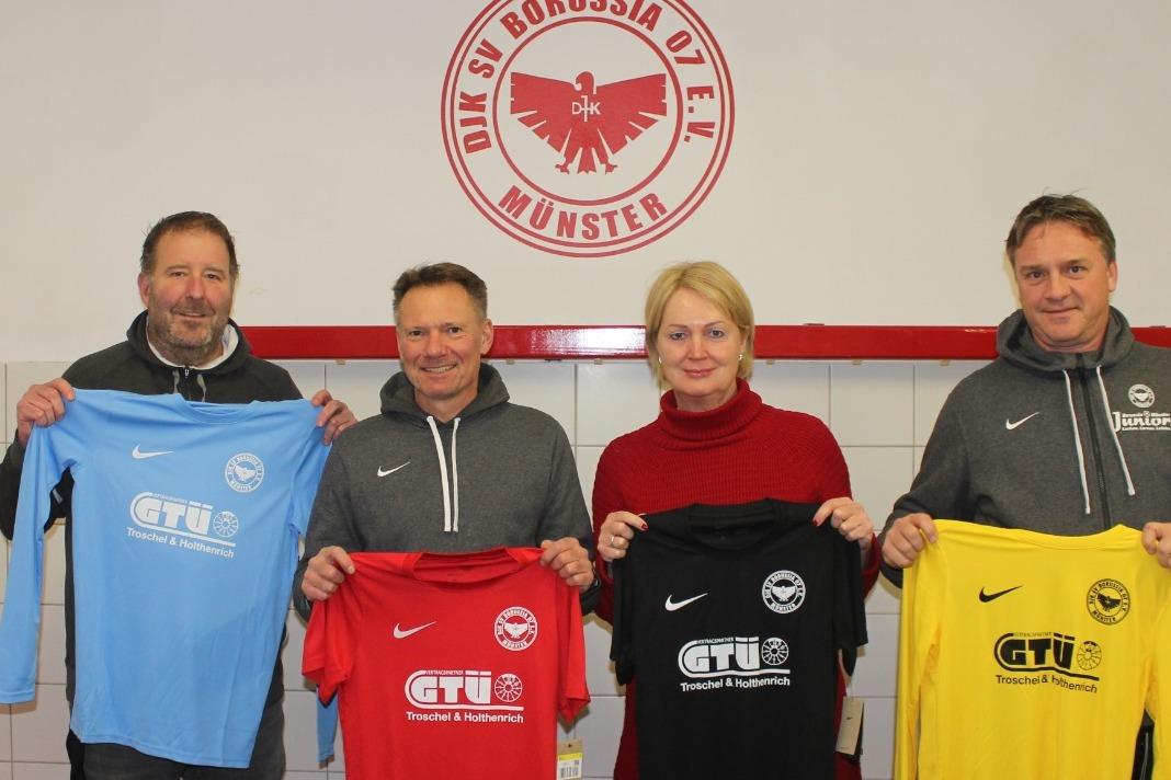 Vier Personen in Sportkleidung pr&auml;sentieren Trikots in verschiedenen Farben vor einer Wand mit dem Logo von Borussia M&uuml;nster.