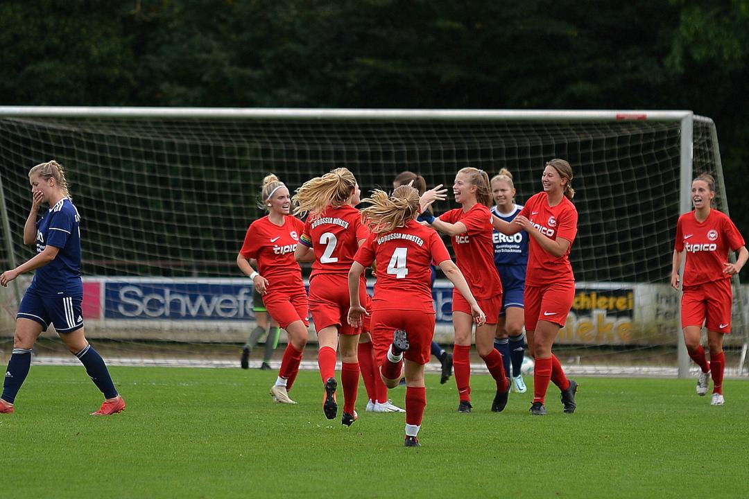 Frauenfu&szlig;ballmannschaft in roten Trikots jubelt nach einem Tor, w&auml;hrend Spielerinnen in blauen Trikots im Hintergrund stehen.