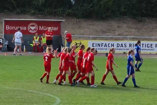 Spieler der Mannschaft in roten Trikots jubeln in einer Gruppenansprache auf einem Fu&szlig;ballplatz, im Hintergrund B&auml;ume und Geb&auml;ude.