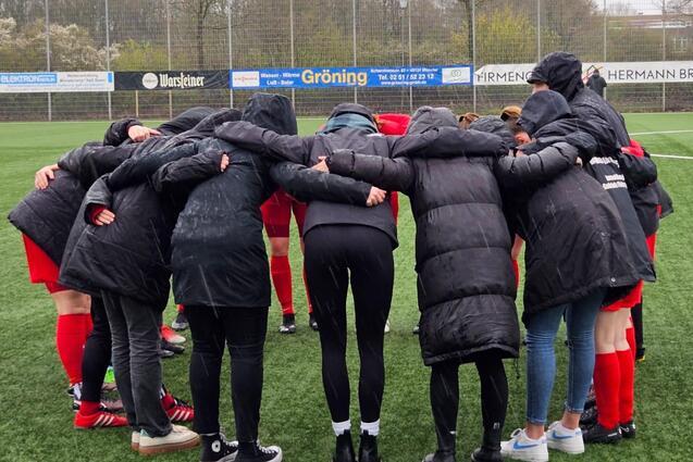 Spielerinnen in schwarzen Jacken bilden einen Kreis auf einem Fu&szlig;ballfeld und versammeln sich f&uuml;r eine Besprechung.