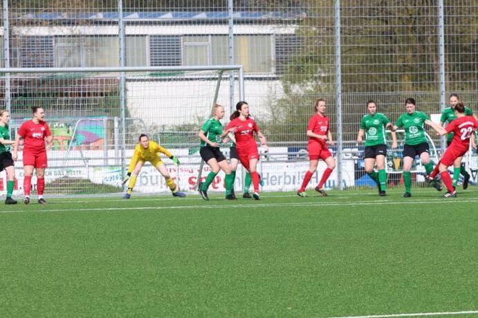 Zwei Damenfu&szlig;ballmannschaften in roten und gr&uuml;nen Trikots spielen auf einem Rasenplatz, Torwart bereitet sich vor.
