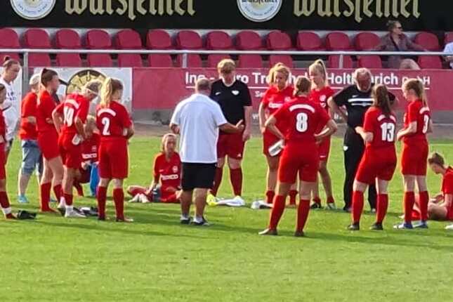 Gruppe von Fu&szlig;ballspielerinnen in roten Trikots um einen Trainer auf einem Rasenplatz versammelt.