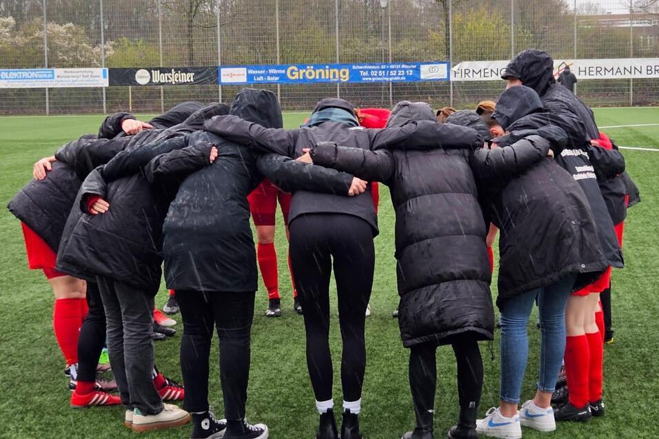 Spielerinnen in schwarzen Jacken bilden einen Kreis auf einem Fu&szlig;ballfeld und versammeln sich f&uuml;r eine Besprechung.