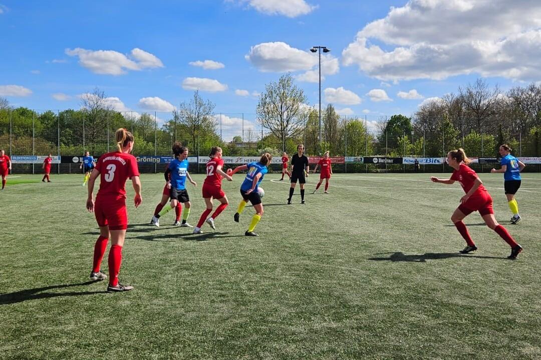 Fu&szlig;ballspiel auf einem Kunstrasenplatz mit Spielern in roten und blauen Trikots unter wolkigem Himmel.