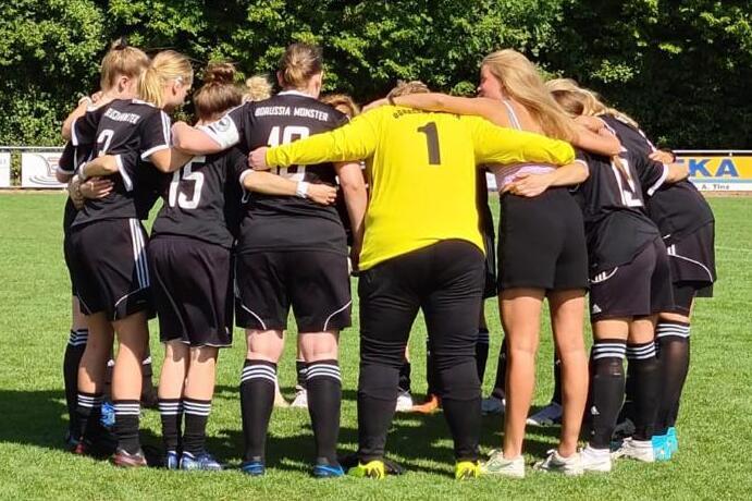 Frauenfu&szlig;ballmannschaft in Schwarz umringt einen Torwart in Gelb auf einem gr&uuml;nen Rasenplatz, im Team-Huddle.