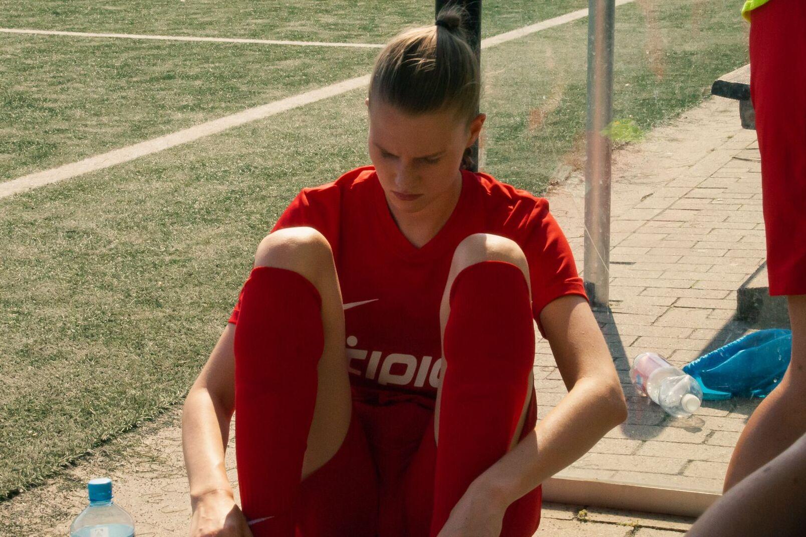 Spielerin in rotem Trikot sitzt auf der Ersatzbank und zieht ihre Socken an, w&auml;hrend sie auf einen Fu&szlig;ballplatz blickt.