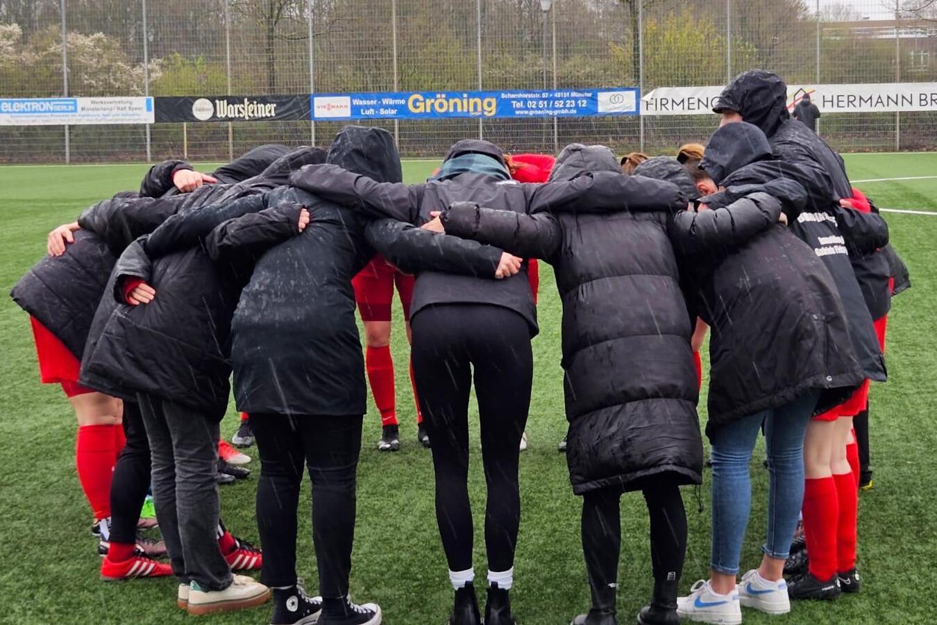 Spielerinnen in schwarzen Jacken bilden einen Kreis auf einem Fu&szlig;ballfeld und versammeln sich f&uuml;r eine Besprechung.