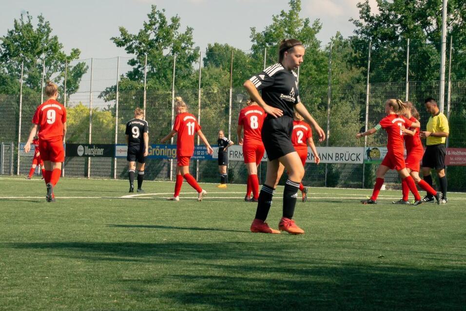 Spielszene auf einem Fu&szlig;ballplatz: Spielerinnen in roten Trikots und Schiedsrichterin auf Rasen; einige Spieler stehen, andere laufen.