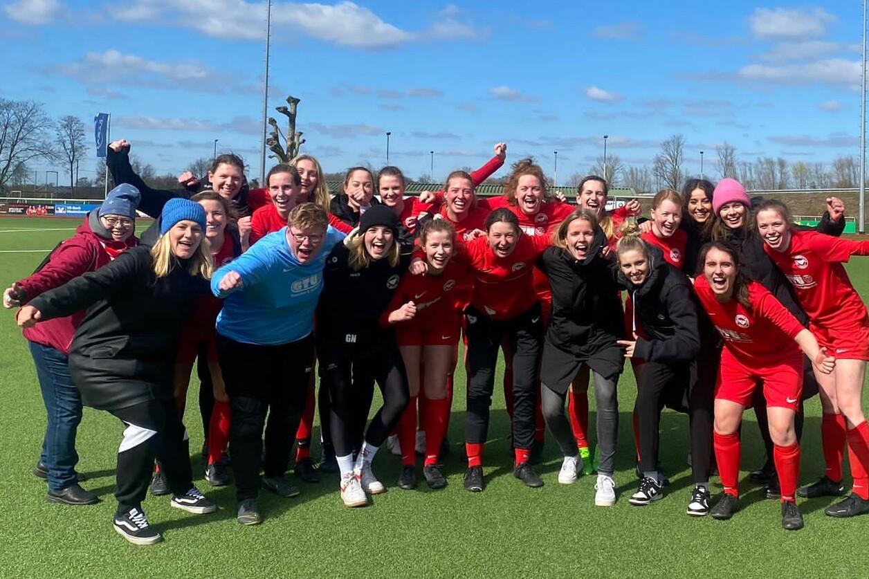 Gruppenshot von Frauen in roten Fu&szlig;balltrikots, feiernd auf einem gr&uuml;nen Kunstrasenfeld unter blauem Himmel.