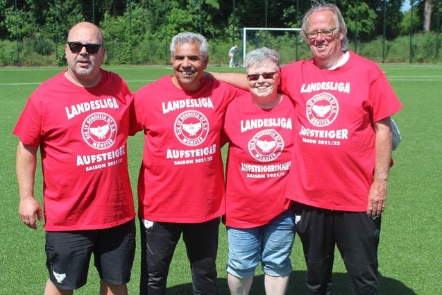 Vier Personen in roten T-Shirts mit dem Aufdruck &bdquo;Landesliga Aufsteiger&ldquo; posieren auf einem gr&uuml;nen Fu&szlig;ballplatz.
