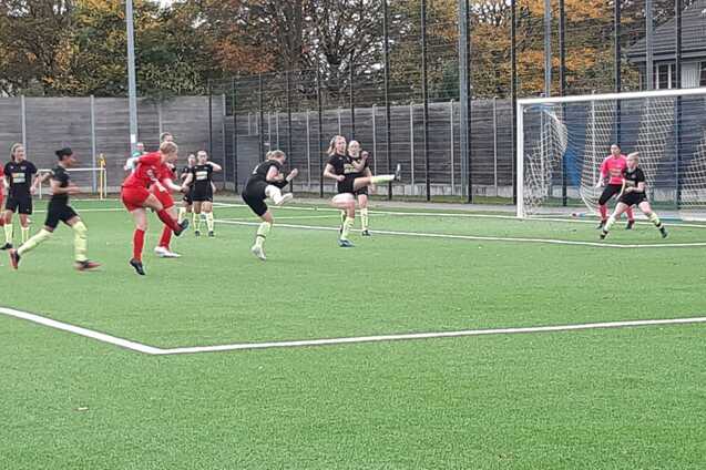 Szenenbild eines Fu&szlig;ballspiels mit mehreren Spielern in roten und schwarzen Trikots auf einem Kunstrasenplatz.