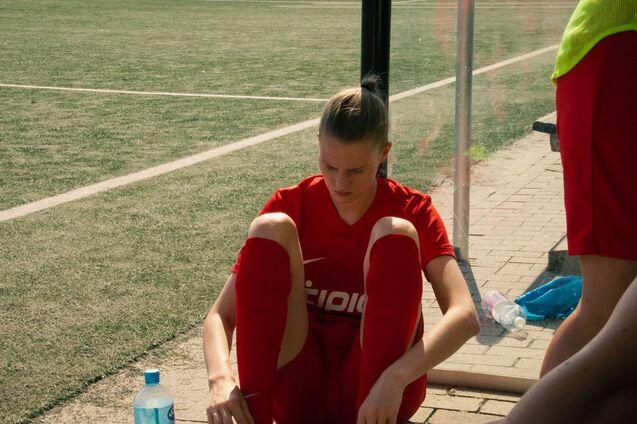 Spielerin in rotem Trikot sitzt auf der Ersatzbank und zieht ihre Socken an, w&auml;hrend sie auf einen Fu&szlig;ballplatz blickt.