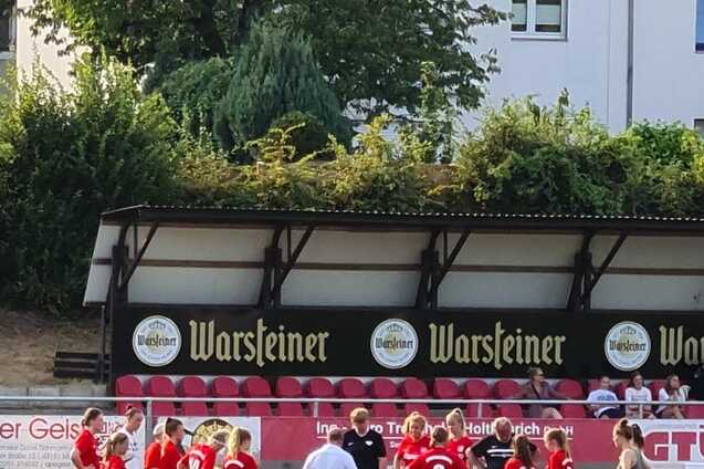 Gruppe von Fu&szlig;ballspielerinnen in roten Trikots um einen Trainer auf einem Rasenplatz versammelt.