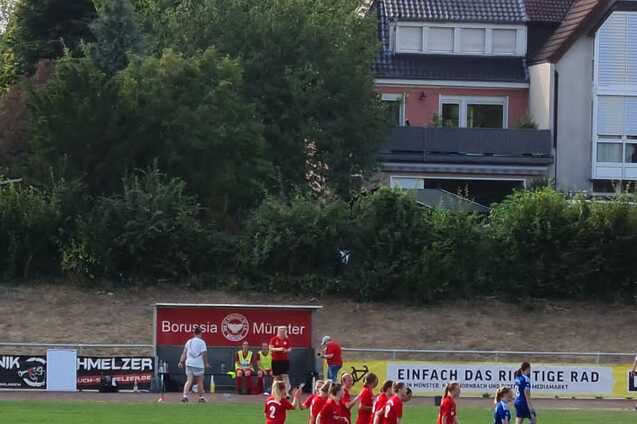 Spieler der Mannschaft in roten Trikots jubeln in einer Gruppenansprache auf einem Fu&szlig;ballplatz, im Hintergrund B&auml;ume und Geb&auml;ude.