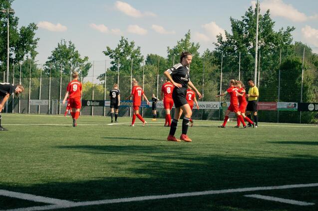 Spielszene auf einem Fu&szlig;ballplatz: Spielerinnen in roten Trikots und Schiedsrichterin auf Rasen; einige Spieler stehen, andere laufen.