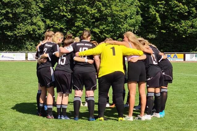 Frauenfu&szlig;ballmannschaft in Schwarz umringt einen Torwart in Gelb auf einem gr&uuml;nen Rasenplatz, im Team-Huddle.