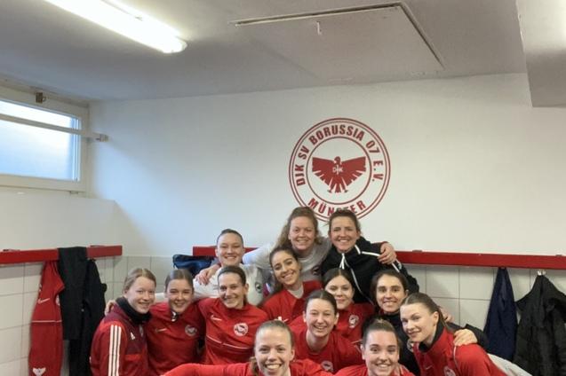 Gruppenshot von zw&ouml;lf Fu&szlig;ballspielerinnen in roten Trikots, l&auml;chelnd im Umkleideraum vor Vereinslogo.
