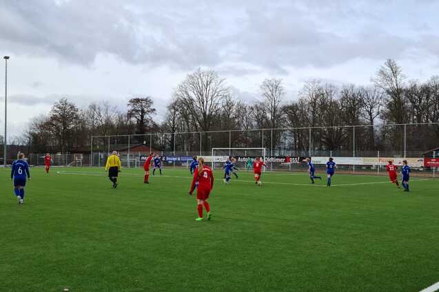 Fu&szlig;ballspiel auf einem Kunstrasenplatz mit Spielern in roten und blauen Trikots und einem Schiedsrichter.