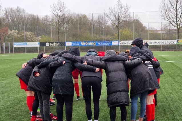 Spielerinnen in schwarzen Jacken bilden einen Kreis auf einem Fu&szlig;ballfeld und versammeln sich f&uuml;r eine Besprechung.