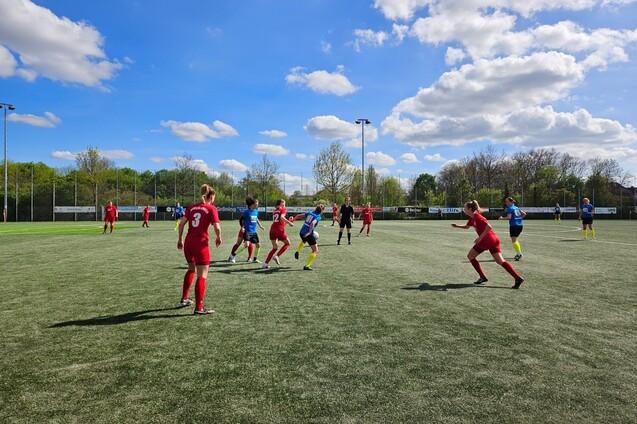 Fu&szlig;ballspiel auf einem Kunstrasenplatz mit Spielern in roten und blauen Trikots unter wolkigem Himmel.