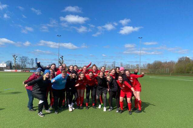 Gruppenshot von Frauen in roten Fu&szlig;balltrikots, feiernd auf einem gr&uuml;nen Kunstrasenfeld unter blauem Himmel.