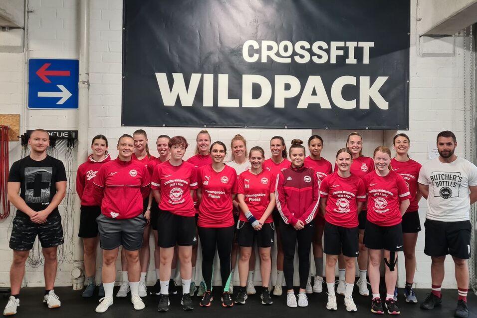Gruppe von CrossFit-Sportlern in roten Shirts posiert vor einer Wand mit dem Schriftzug &bdquo;CROSSFIT WILDPACK&ldquo;.