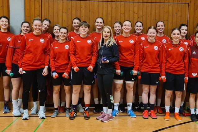 Gruppenfoto von 15 Frauen in roten Trainingsjacken mit Hanteln in der Hand, posing in einer Sporthalle.