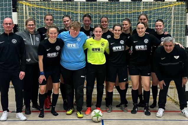 Gruppenfoto eines Frauenteams in schwarzen Trikots vor einem Tor in einer Sporthalle, mit Trainer und Betreuer.
