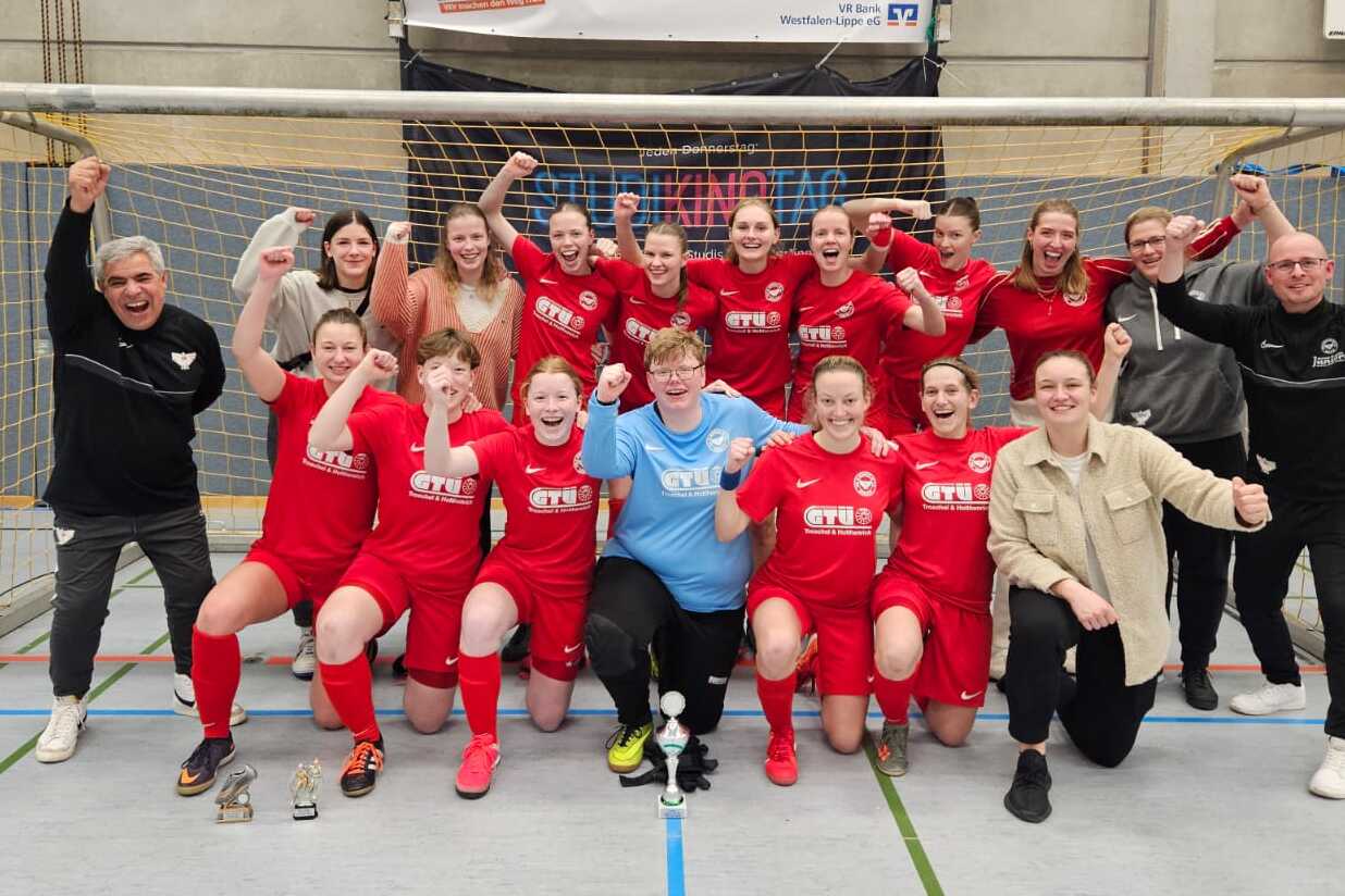 Gruppenshot eines Frauenfu&szlig;ballteams in roten Trikots, jubelnd vor einem Tor mit Pokalen in der Halle.