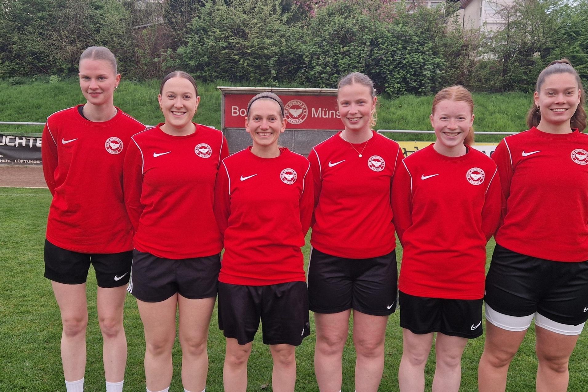 Sechs Frauen stehen in roten Trikots auf einem Fu&szlig;ballplatz, umgeben von gr&uuml;ner Wiese und B&auml;umen im Hintergrund.