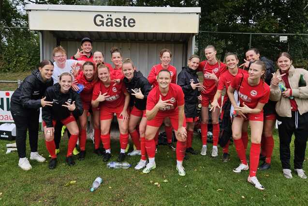 Gruppenselfie einer Fu&szlig;ballmannschaft in roten Trikots, l&auml;chelnd vor der Ersatzbank mit dem Schild &bdquo;G&auml;ste&ldquo;.