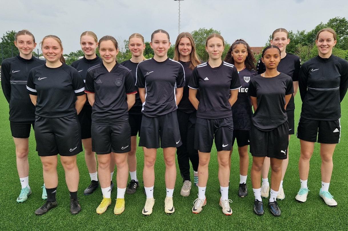 Gruppe von zw&ouml;lf Frauen in schwarzen Sporttrikots steht auf einem Fu&szlig;ballfeld mit Kunstrasen.