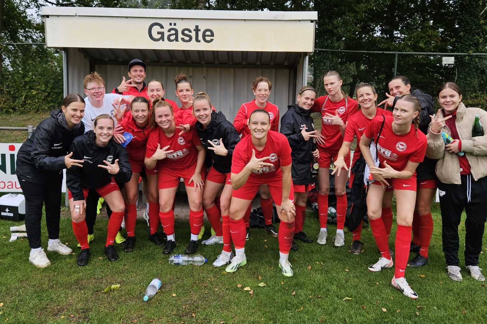 Gruppenselfie einer Fu&szlig;ballmannschaft in roten Trikots, l&auml;chelnd vor der Ersatzbank mit dem Schild &bdquo;G&auml;ste&ldquo;.