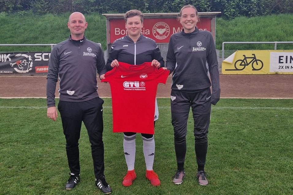 Drei Personen stehen auf einem Fu&szlig;ballplatz und halten ein rotes Trikot mit einem Logo in der Mitte.