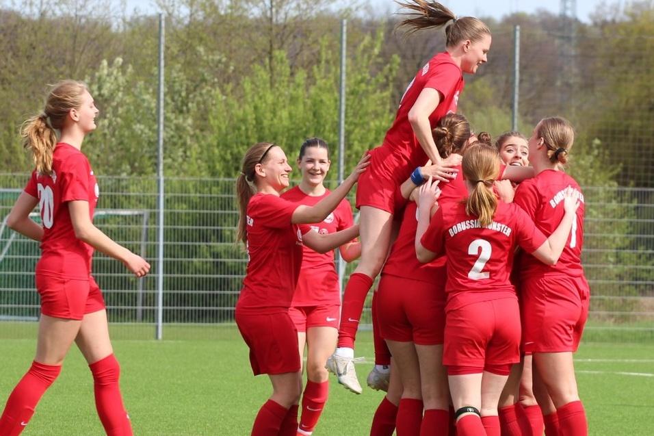 Feiernde Fu&szlig;ballmannschaft in roten Trikots umarmt eine Spielerin auf einem gr&uuml;nen Kunstrasenplatz.