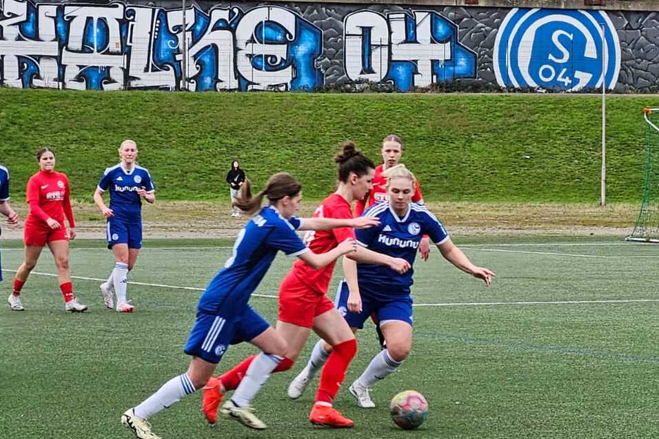 Spielszene auf einem Fu&szlig;ballfeld mit Spielerinnen in blauen und roten Trikots, die um den Ball k&auml;mpfen. Graffiti im Hintergrund.