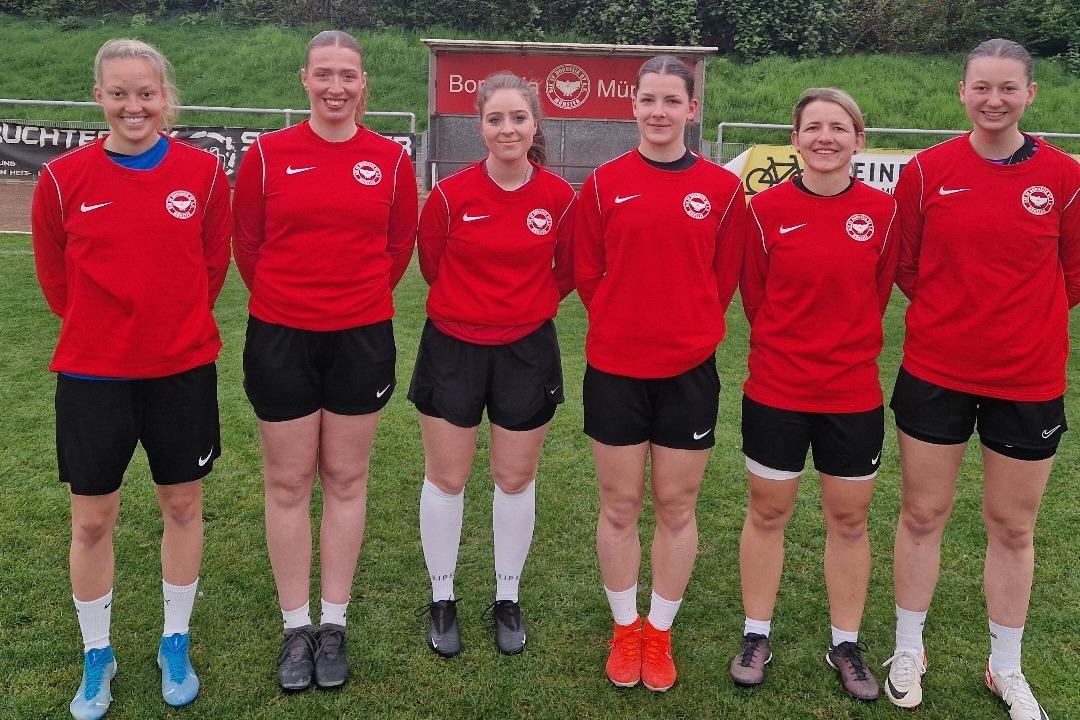 Sechs Fu&szlig;ballerinnen in roten Trikots stehen auf einem gr&uuml;nen Platz, im Hintergrund B&auml;ume und eine Stadionwand.