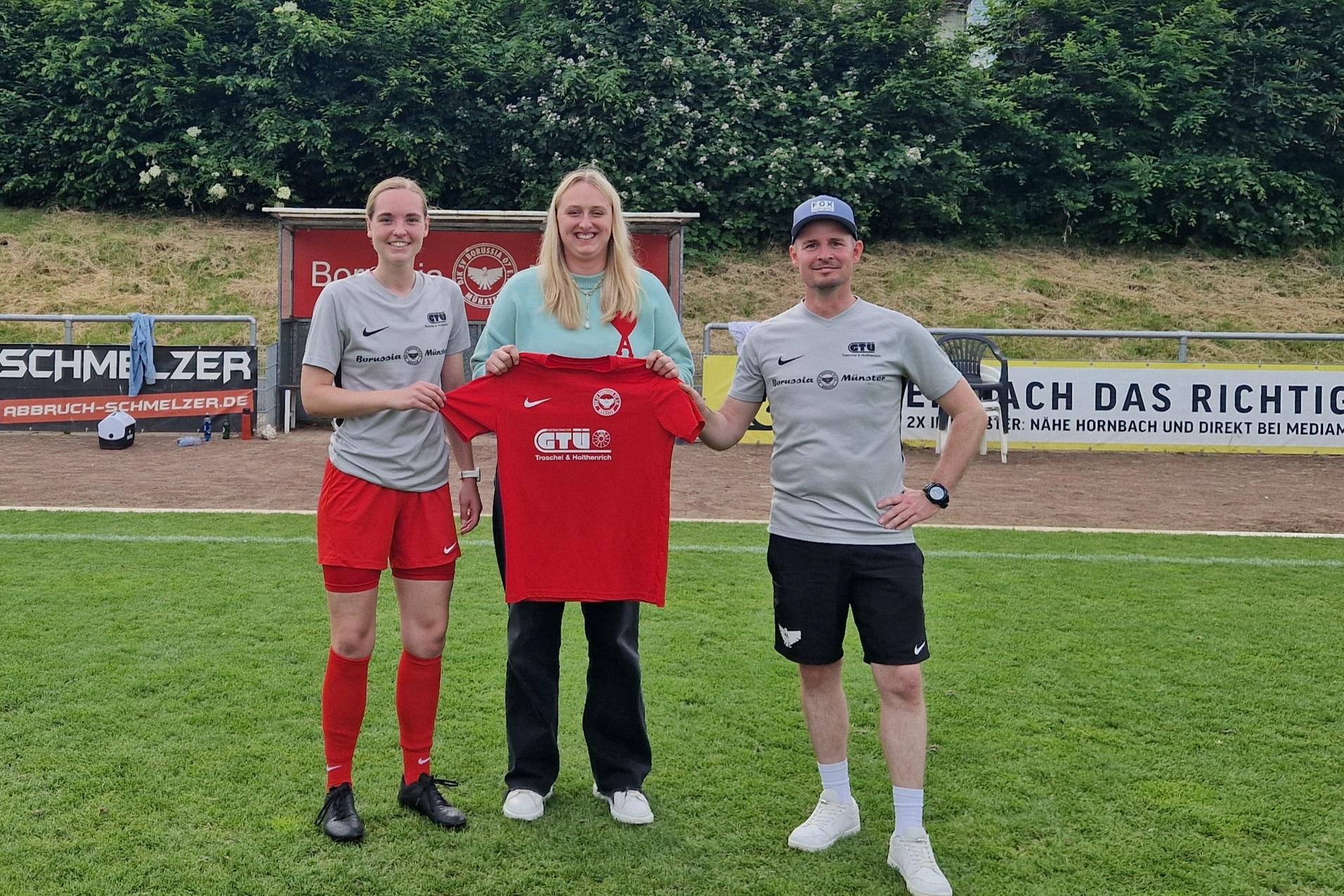 Drei Personen stehen auf einem Fu&szlig;ballfeld, halten ein rotes Trikot und l&auml;cheln in die Kamera. Hintergrund mit B&auml;umen.