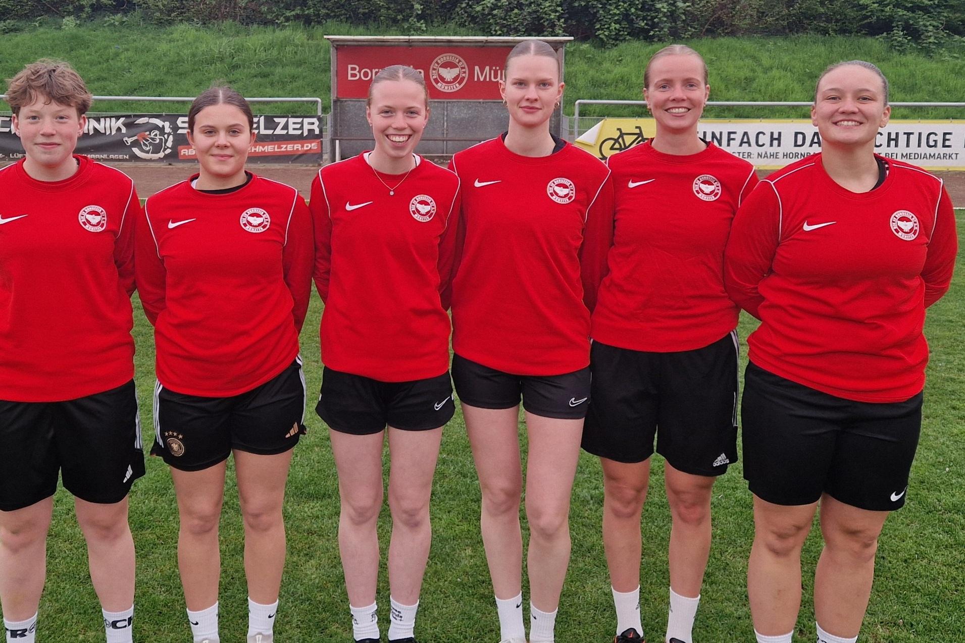 Sechs Fu&szlig;ballspielerinnen in roten Trikots stehen auf einem Sportplatz, im Hintergrund B&auml;ume und eine Werbetafel.