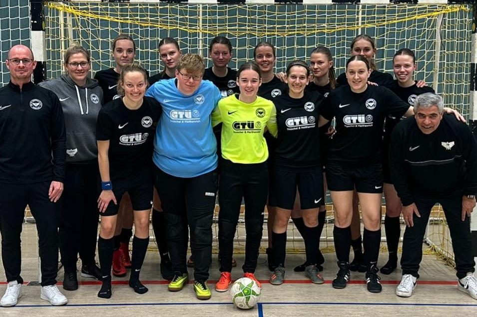 Gruppenfoto eines Frauenteams in schwarzen Trikots vor einem Tor in einer Sporthalle, mit Trainer und Betreuer.