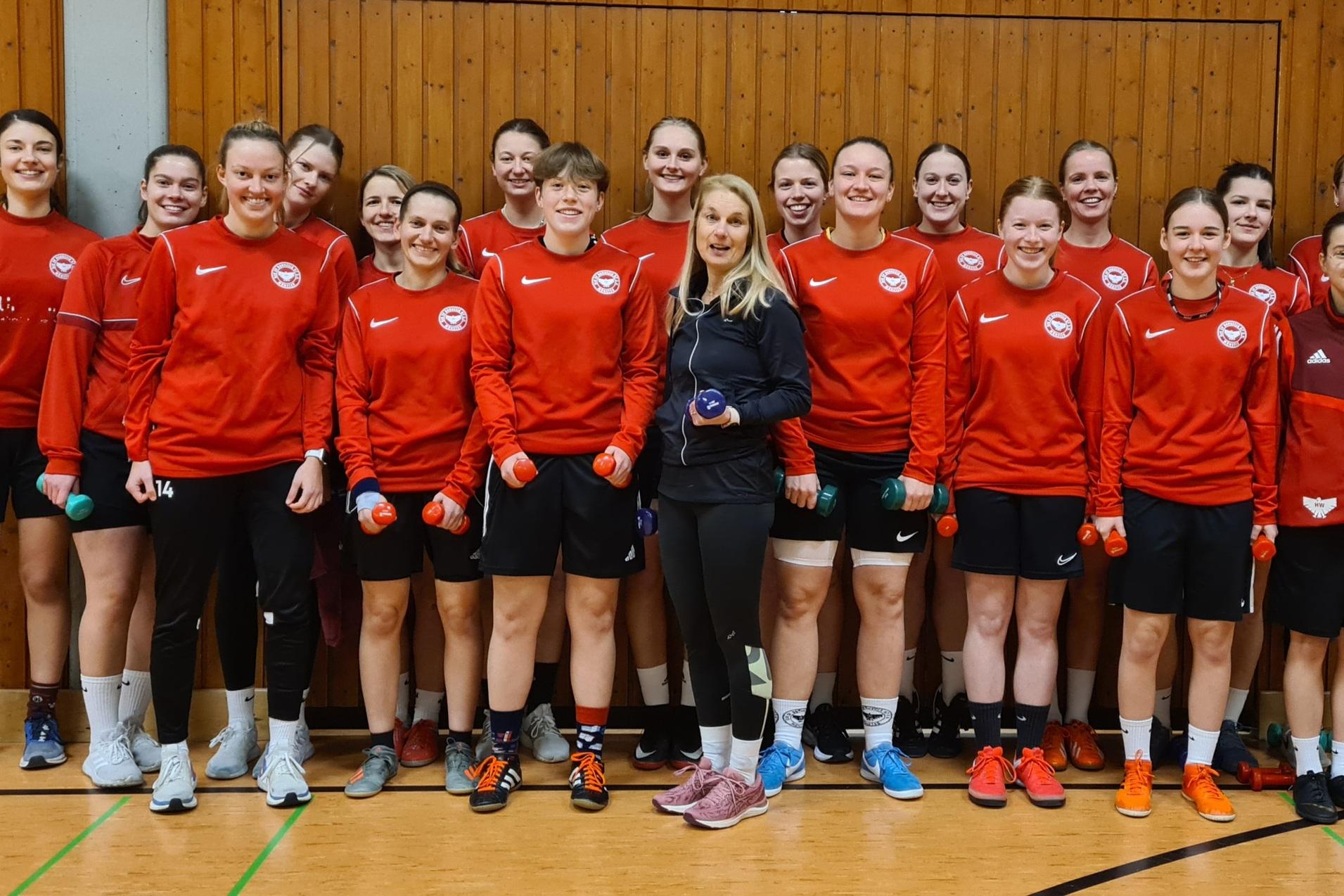 Gruppenfoto von 15 Frauen in roten Trainingsjacken mit Hanteln in der Hand, posing in einer Sporthalle.