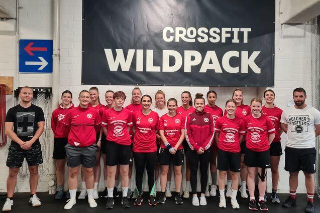 Gruppe von CrossFit-Sportlern in roten Shirts posiert vor einer Wand mit dem Schriftzug &bdquo;CROSSFIT WILDPACK&ldquo;.
