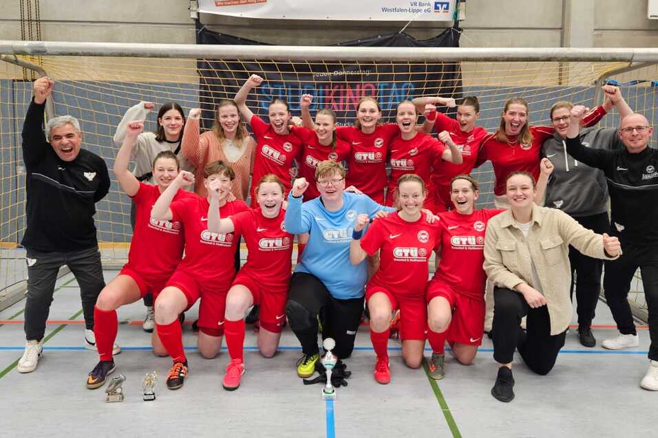 Gruppenshot eines Frauenfu&szlig;ballteams in roten Trikots, jubelnd vor einem Tor mit Pokalen in der Halle.