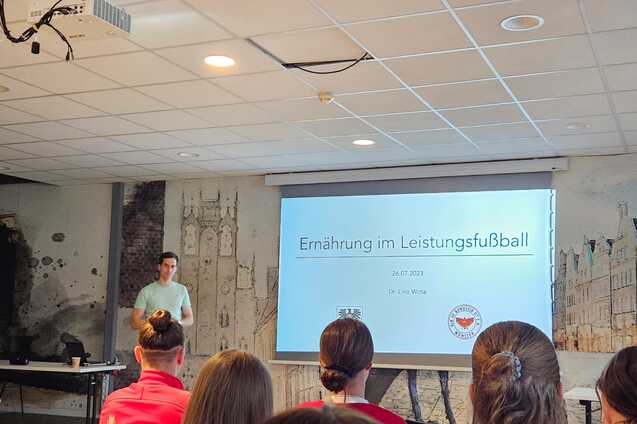 Mann pr&auml;sentiert zum Thema &bdquo;Ern&auml;hrung im Leistungssport&ldquo; vor Publikum in einem modernen Seminarraum.
