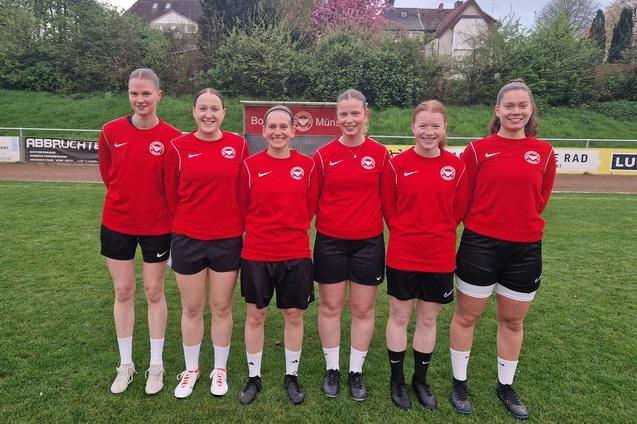 Sechs Frauen stehen in roten Trikots auf einem Fu&szlig;ballplatz, umgeben von gr&uuml;ner Wiese und B&auml;umen im Hintergrund.