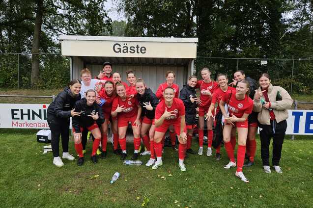 Gruppenselfie einer Fu&szlig;ballmannschaft in roten Trikots, l&auml;chelnd vor der Ersatzbank mit dem Schild &bdquo;G&auml;ste&ldquo;.