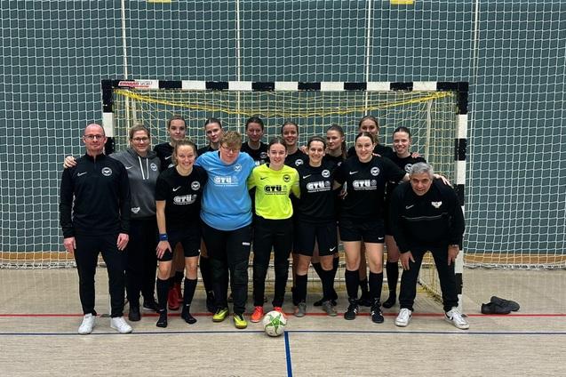 Gruppenfoto eines Frauenteams in schwarzen Trikots vor einem Tor in einer Sporthalle, mit Trainer und Betreuer.