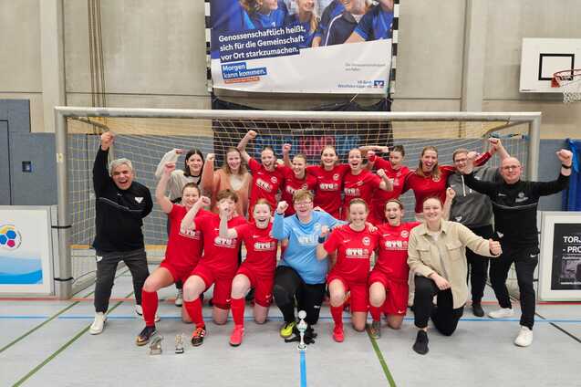 Gruppenshot eines Frauenfu&szlig;ballteams in roten Trikots, jubelnd vor einem Tor mit Pokalen in der Halle.