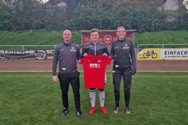 Drei Personen stehen auf einem Fu&szlig;ballplatz und halten ein rotes Trikot mit einem Logo in der Mitte.
